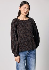 Top marrone a maniche lunghe con stampa leopardata nera, caratterizzato da un colletto rotondo e polsini arricciati, abbinato a jeans azzurri chiari.