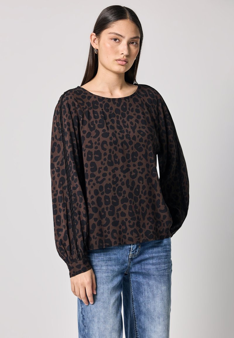 Top marrone a maniche lunghe con stampa leopardata nera, caratterizzato da un colletto rotondo e polsini arricciati, abbinato a jeans azzurri chiari.