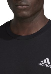 Detailní záběr na osobu ve černém tričku Adidas s bílým logem na hrudi, zaměřující se na krk, rty a horní část trupu.