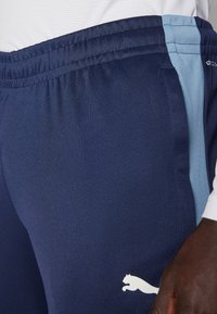 Calças de desporto na cor azul-marinho com um painel lateral azul-claro, cintura elástica e um logótipo de gato branco. O tecido parece suave e texturizado.