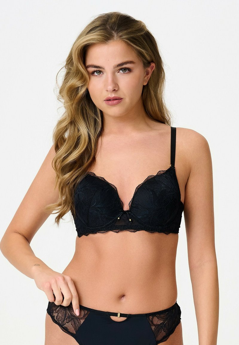 Femme aux longs cheveux ondulés portant un soutien-gorge en dentelle noire et une culotte assortie, posant devant un fond clair uni.