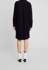 Abito camicia in corda di colore blu scuro, con maniche lunghe, polsini con bottoni, uno spacco sul retro per la libertà di movimento, orlo curvo, abbinato a stivali bianchi con tacco.