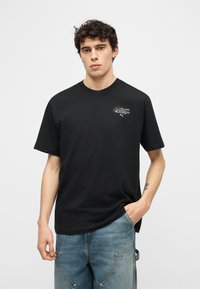 Νεαρός με σγουρά μαλλιά που φοράει μαύρο μπλουζάκι με τύπωμα και φαρδιά μπλε τζιν, στέκεται μπροστά σε απλό ανοιχτό φόντο.