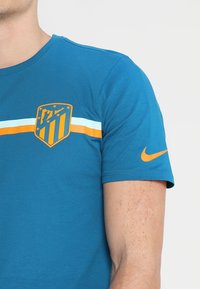 Camiseta de algodón azul con el logo del Atlético de Madrid en color naranja, con rayas horizontales blancas y naranjas en el pecho y un logo de Nike en la manga.