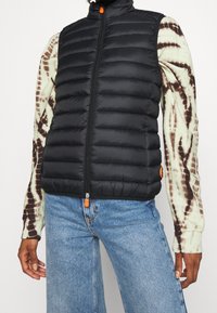 Sort dunvest med lodret quiltning, høj krave, frontlynlås og orange lynlåstrækker. Båret over en tie-dye langærmet skjorte.