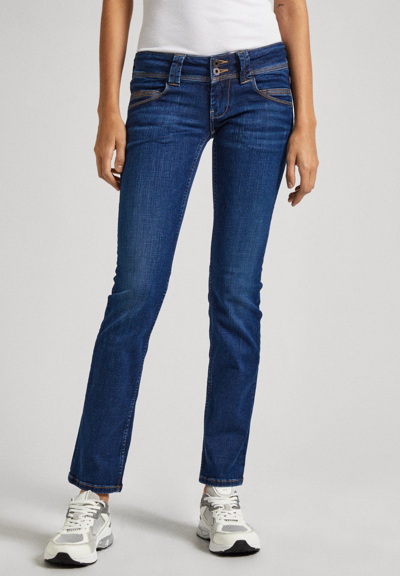 Slim-fit blue denim jeans with a mid-rise waist, five-pocket design, and contrasting stitching. Paired with white sneakers.