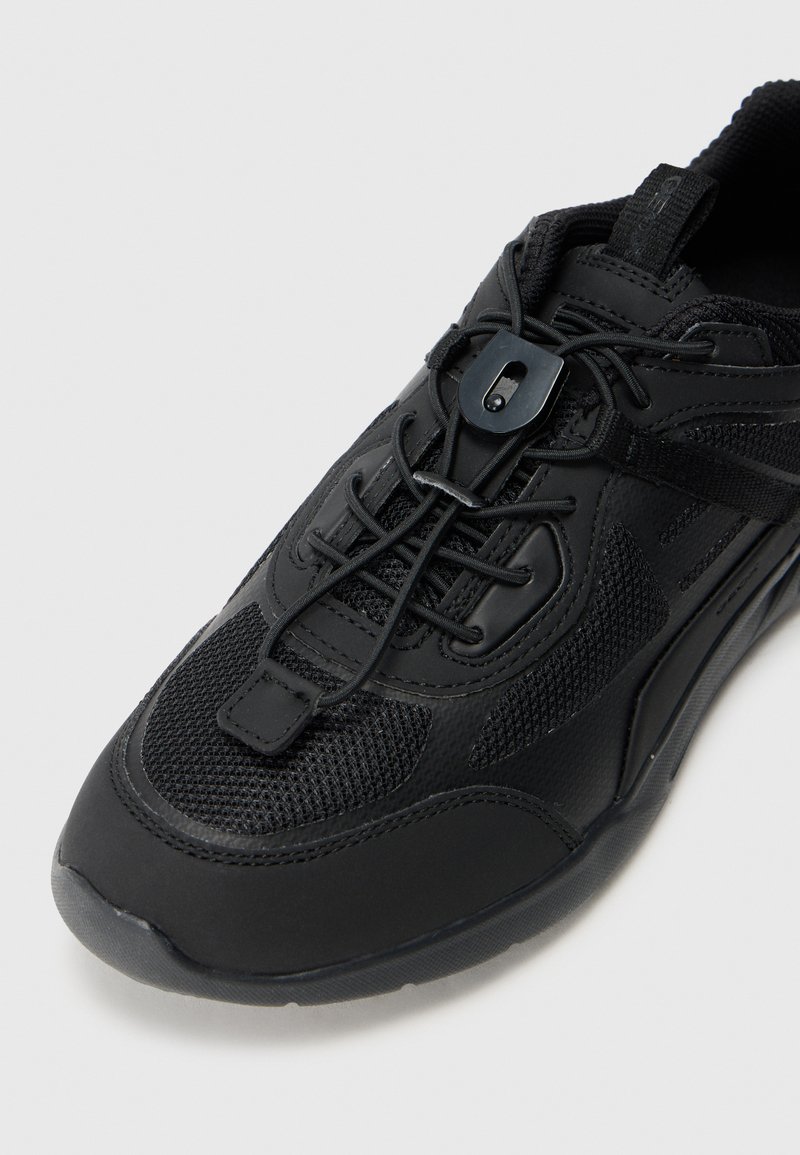 Czarne buty sportowe z siateczkowych i syntetycznych paneli, elastyczne sznurowadła bez wiązania zabezpieczone plastikowym zaciskiem oraz pętelka na języku.