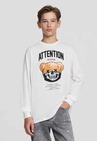 Biała koszulka z długim rękawem z grafiką niedźwiedzia ze czaszką, w towarzystwie czarnego napisu "POSZUKIWACZ UWAGI". Szare zniszczone dżinsy.