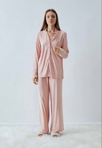 Kvinna som står på en vit fluffig matta iklädd ljusrosa pyjamas och beige tofflor framför en vit vägg.