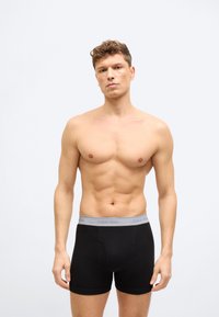 Άνδρας χωρίς πουκάμισο με κοντά σγουρά μαλλιά, φορώντας μαύρα μποξέρ Calvin Klein με γκρι μέση, στέκεται μπροστά σε απλό φωτεινό φόντο.