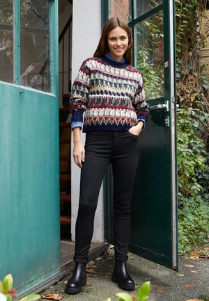 Πλεχτό πουλόβερ σε πολύχρωμα σχέδια με ναυ blå, κόκκινο και κρεμ απόχρωση. Συνδυασμένο με εφαρμοστά μαύρα τζιν και μαύρες μπότες μέχρι τον αστράγαλο, στέκεται δίπλα σε μια πράσινη πόρτα.