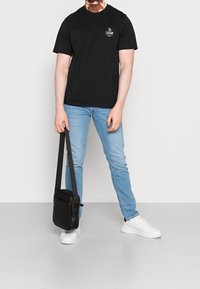 Černé tričko s krátkým rukávem a malým logem, světle modré džíny a bílé tenisky. Z ruky visí černá taška přes rameno se popruhem.