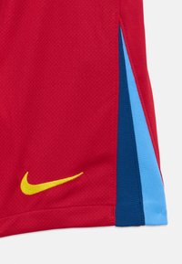 Κόκκινη αθλητική φανέλα με υφή στην επιφάνεια. Διαθέτει κίτρινο λογότυπο Nike και πλαϊνές λεπτομέρειες σε μπλε και ανοιχτό μπλε.