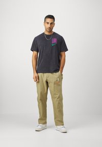 Black short-sleeve T-shirt with purple and green logo accents, paired with beige pants featuring snap buttons and Nike branding, worn with white sneakers.