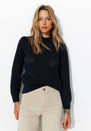 Pull en tricot bleu marine avec un motif en losange texturé, col et poignets côtelés, associé à un pantalon beige clair. Design simple et décontracté.