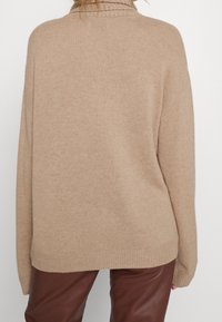 Beige oversized trui met geribbelde hals, gestructureerde breiwerk en verlaagde schouders. Draagt over bruin leren broeken, met een ontspannen zoom.