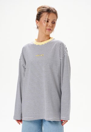 Vrouw draagt een losse zwart-wit gestreepte longsleeve met een gele kraag, gecombineerd met lichtblauwe spijkerbroek, kijkt naar beneden.