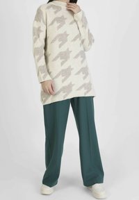 Personne portant un pull oversize beige à motif pied-de-poule, un pantalon large couleur sarcelle et des baskets blanches, debout devant un fond clair uni.