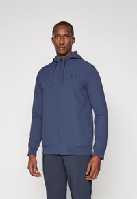 Haut à capuche zippé bleu marine en matériau texturé, doté d'une capuche, de cordons de serrage ajustables et d'un petit logo sur la poitrine.