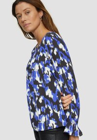Zwarte blouse met een blauw en witte abstracte patroon, lange mouwen en ruches aan de manchetten. Gemaakt van lichtgewicht stof, met een ronde halslijn.