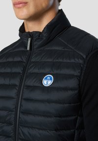 Chaleco acolchado negro con textura acolchada, cuello alto y cremallera frontal. Presenta un logo bordado circular en azul y blanco en el pecho.