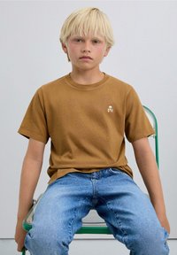 Brown short-sleeve T-shirt with a small white skull logo on the left chest, worn with faded blue jeans, sitting on a green metal chair.