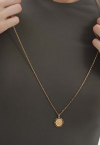 Collar de colgante de oro sobre una cadena delgada, con un charm redondo que presenta un diseño intrincado. Lleva puesto sobre una prenda oscura y texturizada.