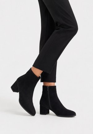 Piernas con pantalones negros ajustados y botines de ante negro con tacones gruesos y cremalleras laterales sobre un fondo blanco.