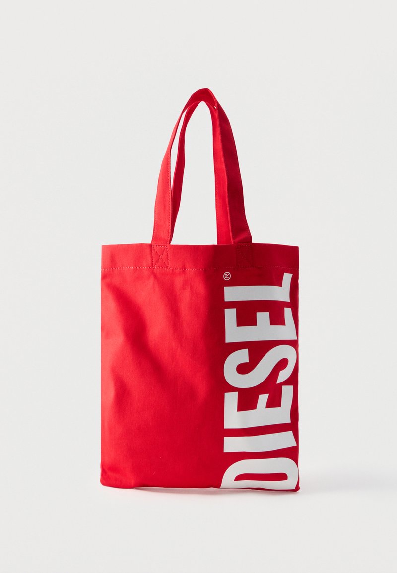 Τσάντα tote από κόκκινο ύφασμα με δύο λαβές, με λευκή κάθετη επιγραφή «DIESEL» στη δεξιά πλευρά πάνω σε απλό φόντο.