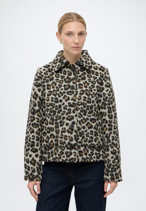 Kvinde med blondt hår iført en leopardprintjakke med knapper og mørke bukser, stående mod en ensfarvet hvid baggrund.