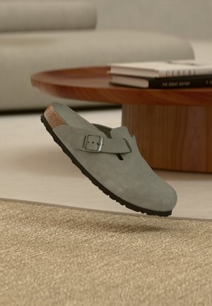 Sabot Birkenstock en daim gris avec sangle à boucle flottant au-dessus d'un tapis beige tissé, près d'une table basse en bois avec des livres empilés.