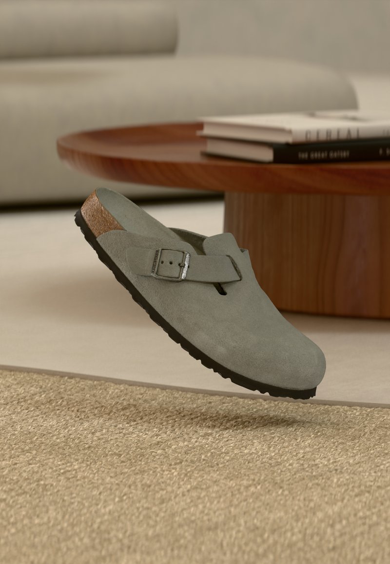 Grå ruskinds Birkenstock-træsko med spænde, der svæver over et beige vævet tæppe, nær et sofabord i træ med stablede bøger.