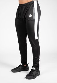 Pantalons de sport noirs avec une coupe ajustée, dotés d'une bande latérale blanche et d'une poche zippée, fabriqués dans un matériau lisse et extensible.