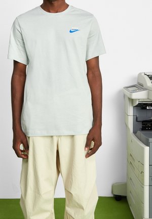 Hellgrünes Baumwoll-T-Shirt mit einem blauen Nike-Logo auf der linken Brust, kombiniert mit creamfarbenen, locker sitzenden Hosen.