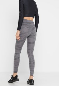 Leggings grigi con motivo a fantasia e design testurizzato, vita alta e scarpe sportive nere. La vista posteriore mostra la vestibilità e il materiale.