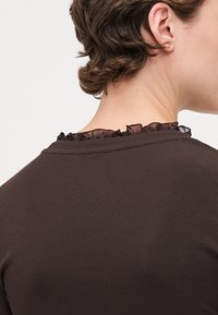 Brunt ribbet topp med rund halslinning som har svart volangkant. Stoffet ser mykt og litt elastisk ut.
