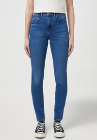 Jeans de mezclilla azul ajustados con cinco bolsillos, ligeras decoloraciones y costuras en contraste, combinados con zapatillas negras de caña alta.