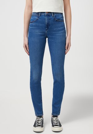 Jeans in denim blu slim-fit con cinque tasche, leggermente sbiaditi e con cuciture a contrasto, abbinati a sneakers alte nere.