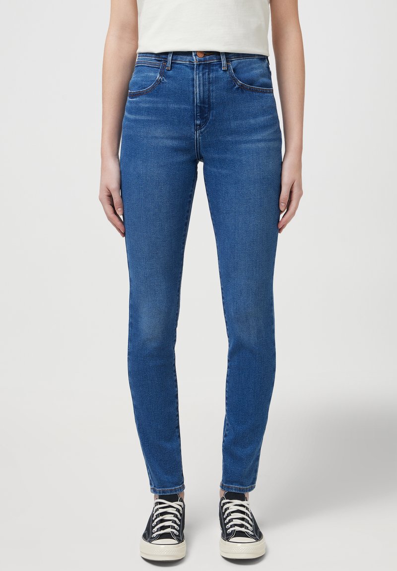 Jeans de mezclilla azul ajustados con cinco bolsillos, ligeras decoloraciones y costuras en contraste, combinados con zapatillas negras de caña alta.