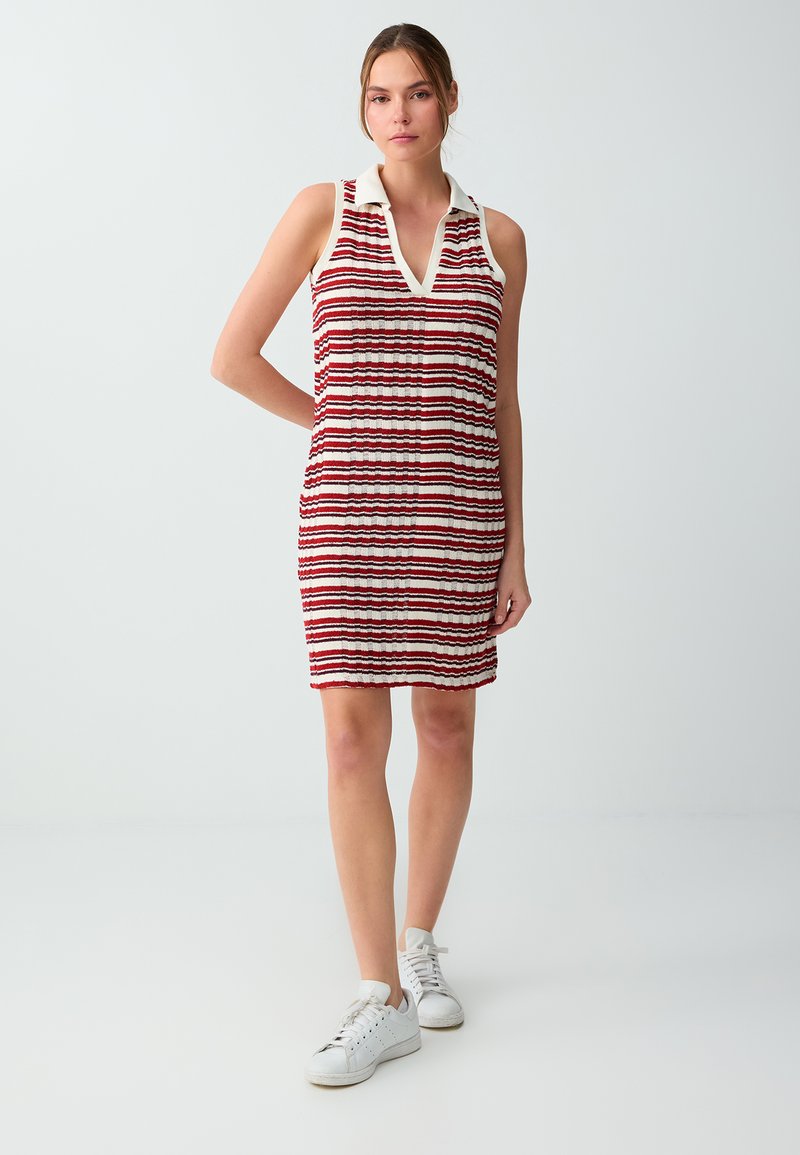 Femme portant une robe rayée rouge, blanche et noire sans manches avec col, debout et portant des baskets blanches sur un fond clair uni.