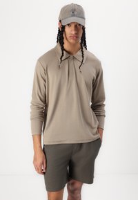 Polo shirt à manches longues beige avec un col, accompagné de shorts vert olive et d'une casquette assortie. Le tissu semble lisse avec une coupe décontractée.