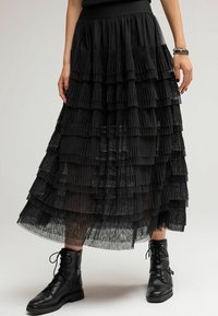 Jupe plissée noire avec des volants superposés, dotée d'une ceinture lisse. Associée à des bottines noires à lacets. Superposition en tissu translucide.