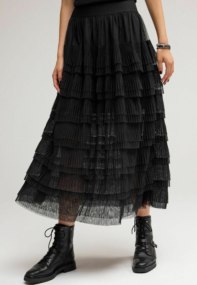 Jupe plissée noire avec des volants superposés, dotée d'une ceinture lisse. Associée à des bottines noires à lacets. Superposition en tissu translucide.