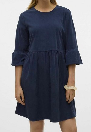Vrouw die een marineblauwe corduroy jurk draagt met 3/4 klokmouwen en een geplooide taille, houdt een doorschijnende beige armband om haar rechterpols.