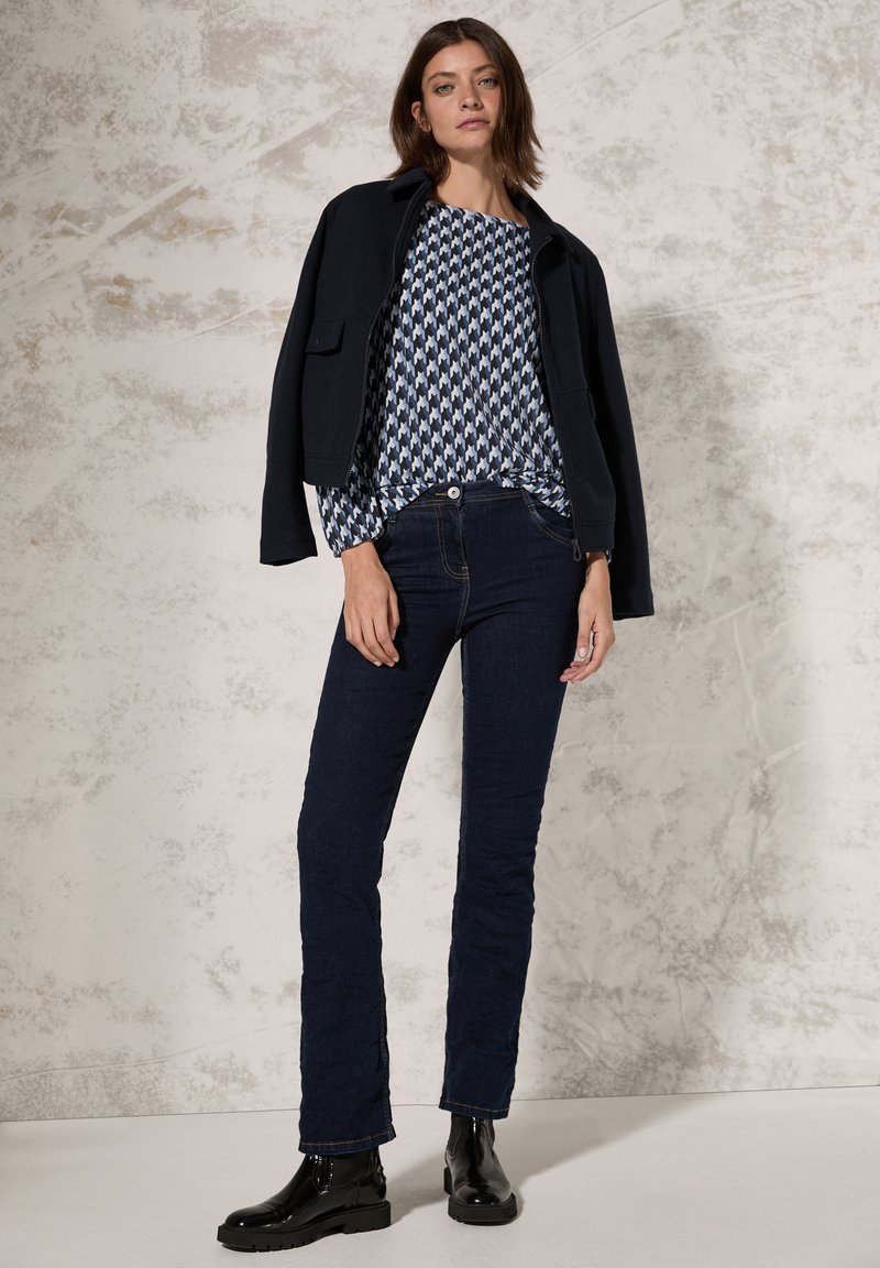 Veste en mélange de laine marine sur un haut à motifs bleu et blanc, associée à un jean en denim foncé et des bottines noires. Textures lisses et coupes ajustées.