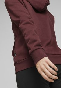 Felpa bordeaux con finitura testurizzata, dotata di un ampio cappuccio e polsini a coste, arricchita da un piccolo etichetta con il marchio nella parte inferiore.