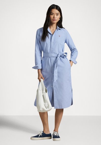 Ung kvinne if�ørt lys blå skjortekjole med belte og marineblå joggesko, holder hvit håndveske med den ene hånden i lommen foran en ensfarget bakgrunn.