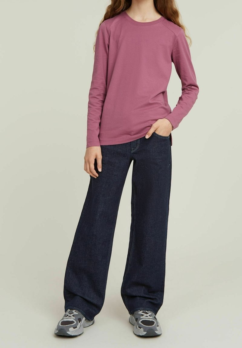 T-shirt mauve à manches longues avec un col rond, associé à un jean large bleu foncé. Le modèle porte des baskets grises avec un design à motifs.