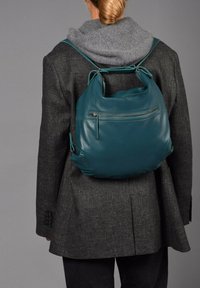 Mochila de cuero verde azulado con forma redondeada, bolsillo delantero con cremallera y correas ajustables, llevada sobre un blazer gris y un suéter.