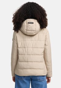 Persoon met krullend haar die een beige gewatteerde pufferjack met capuchon en blauwe jeans draagt, gezien vanaf de achterkant tegen een eenvoudige achtergrond.